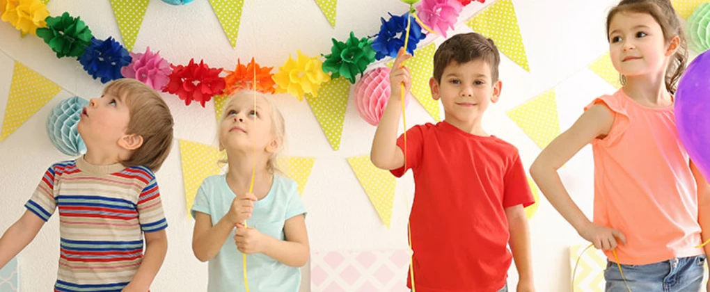 Vier Kinder feiern in einem bunt geschmückten Raum mit Luftballons.