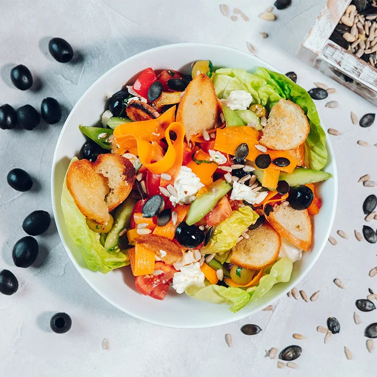 Bowl with a colorful Greek salad of romaine lettuce, tomatoes, cucumbers, carrots, olives, feta, croutons and seeds.
