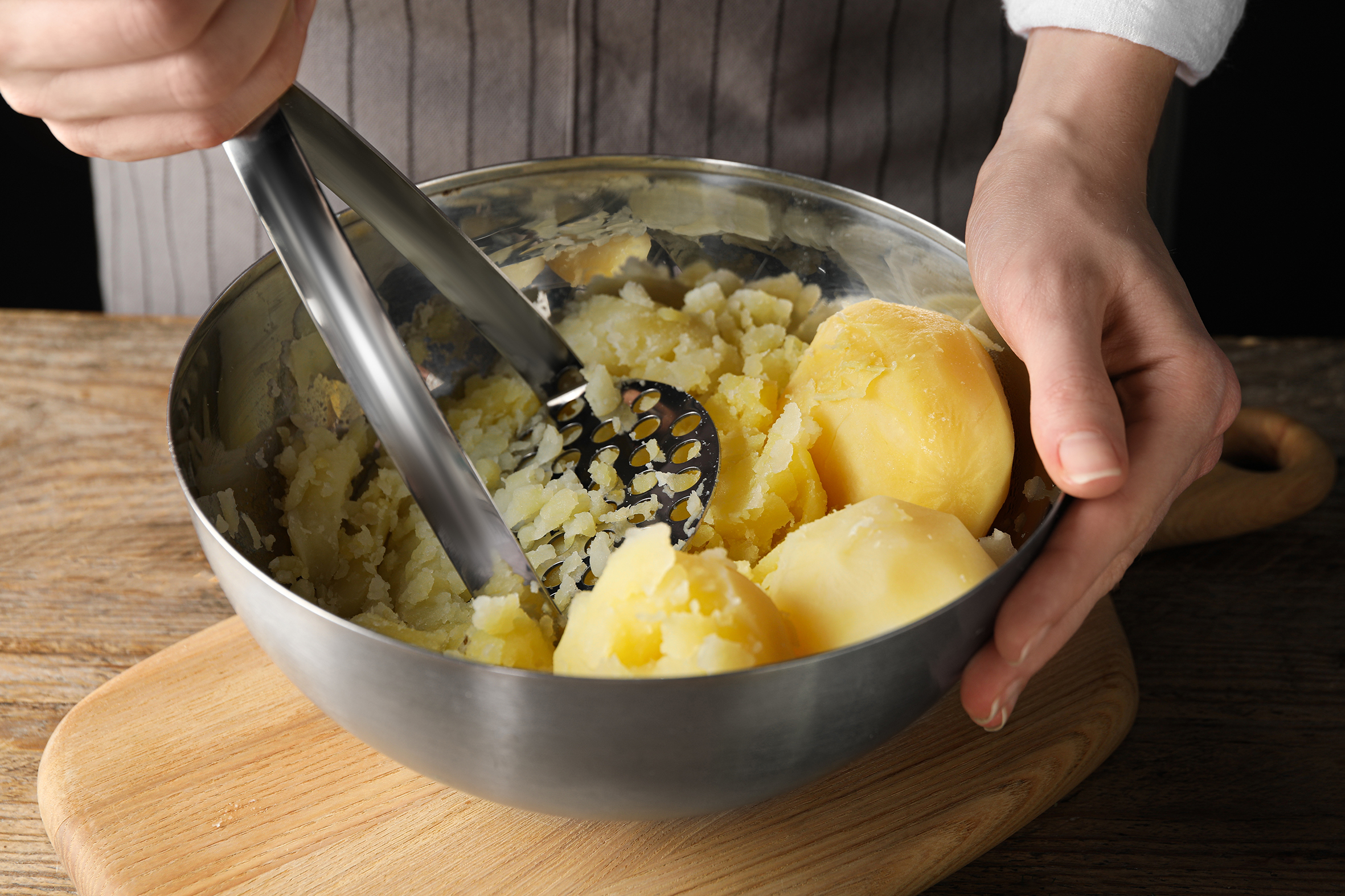 Person zerstampft gekochte Kartoffeln in einer Metallschüssel mit einem Kartoffelstampfer.