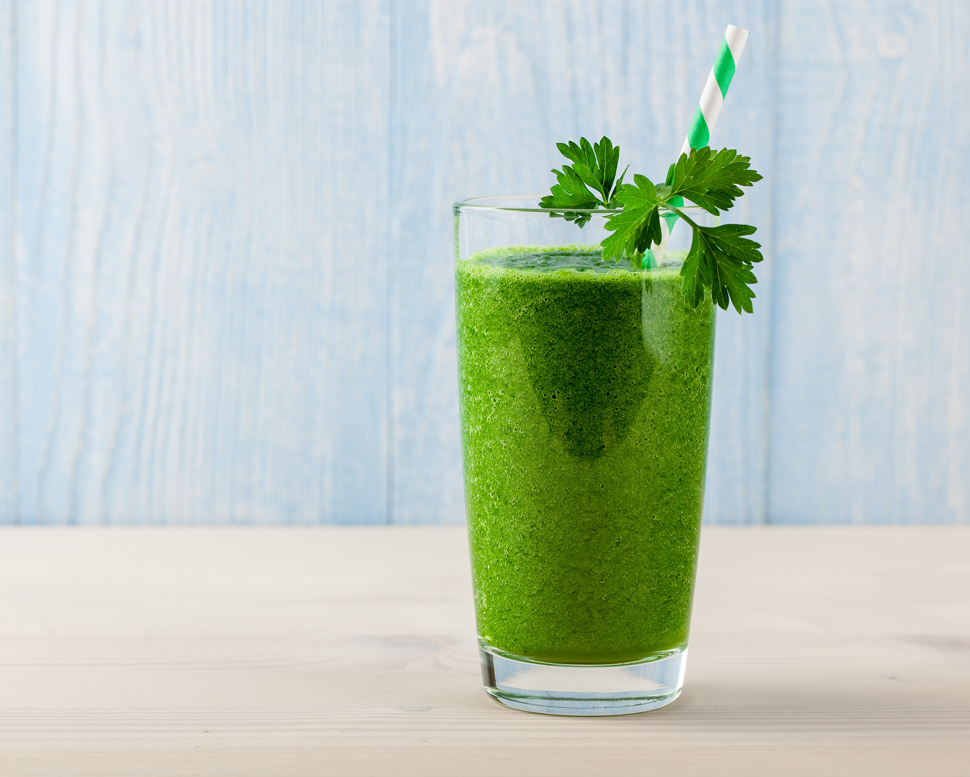Glas mit grünem Petersilien-Smoothie und Trinkhalm, dekoriert mit einem Petersilienblatt, vor hellblauem Holz-Hintergrund.
