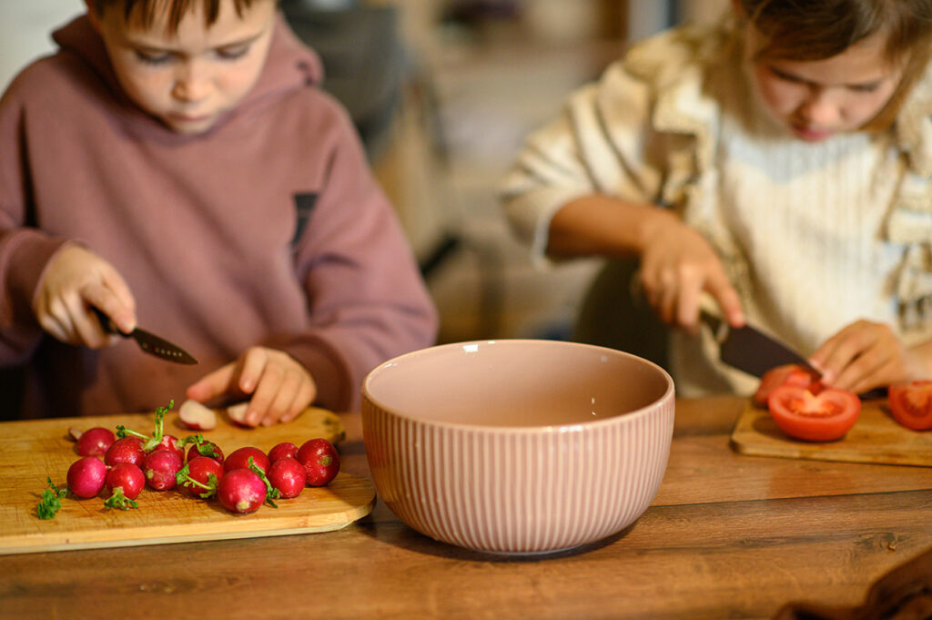 Zwei Kinder schneiden Radieschen und Tomaten auf Holzbrettchen, daneben steht eine große rosa Schüssel.
