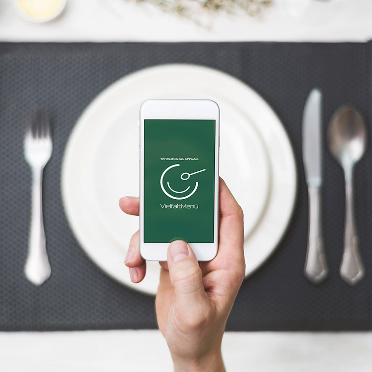 Eine Hand hält ein Smartphone mit der VielfaltMenü-App, das vor einem gedeckten Tisch in einem Restaurant präsentiert wird.