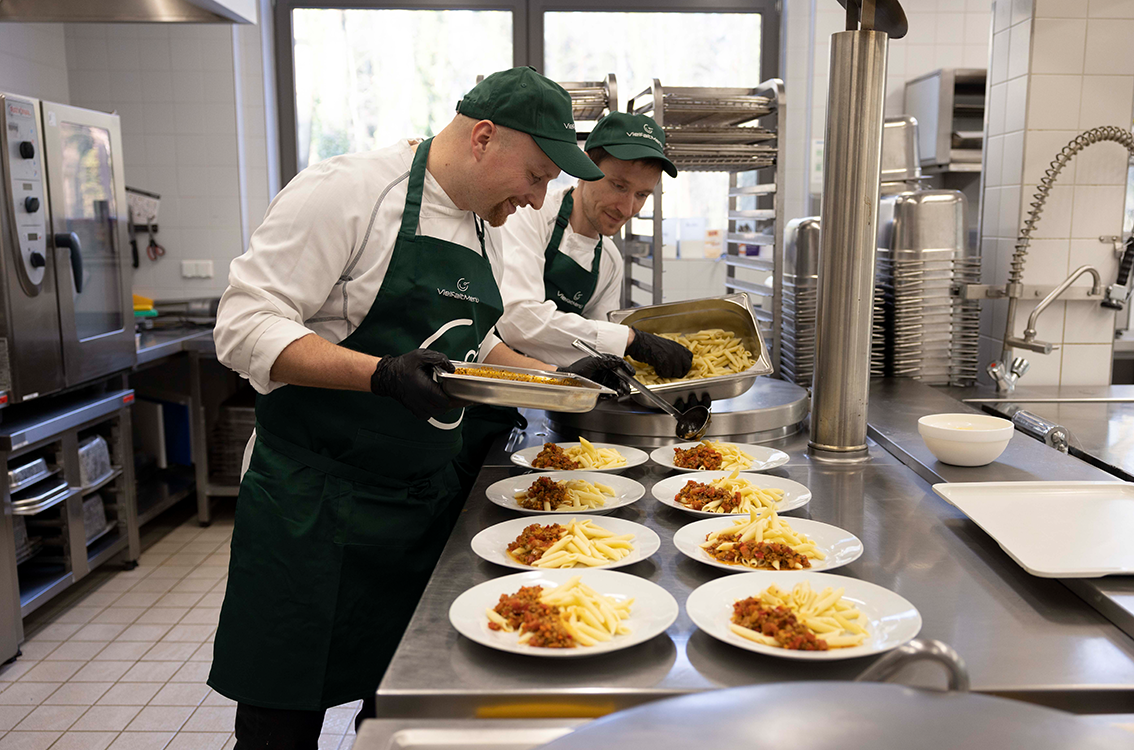 Mitarbeiterinnen und Mitarbeiter von VielfaltMenü portionieren warme Mahlzeiten in einer Großküche auf mehrere Teller.