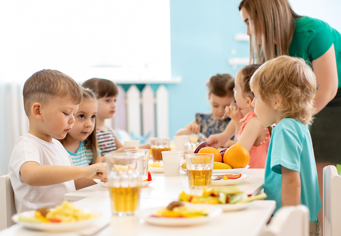 Kinder sitzen gemeinsam an einem Tisch in einer Kita und essen Obst und warme Speisen, eine Fachkraft begleitet die Situation.