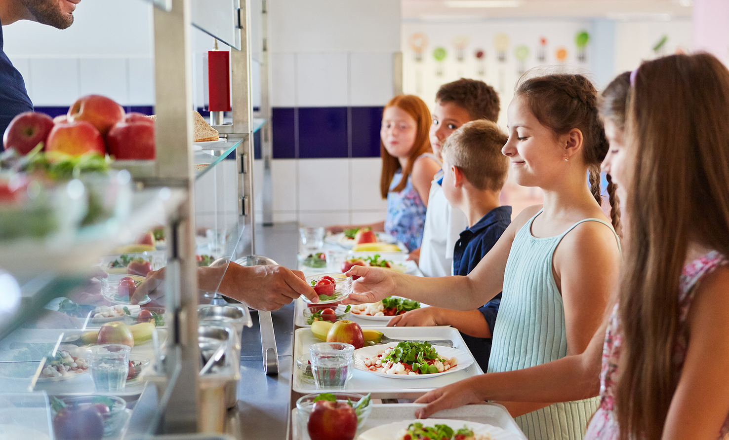 Kinder stehen in einer Schulkantine an und erhalten ein Tablett mit frischem Essen und Obst.