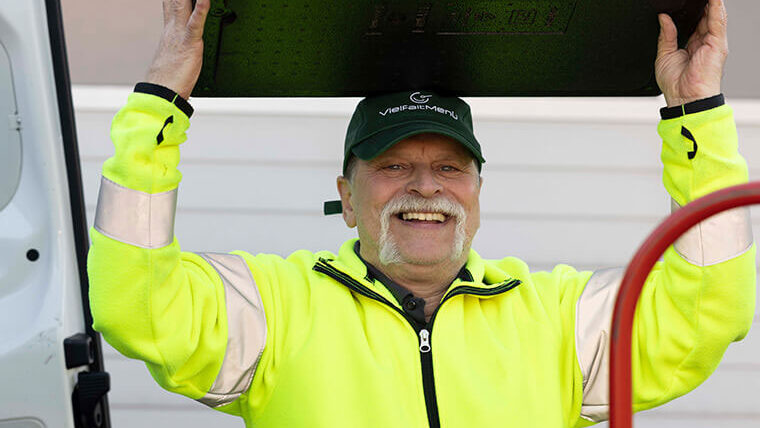 Ein lächelnder Fahrer in einer neongelben Jacke hebt eine schwarze Transportbox über seinen Kopf.