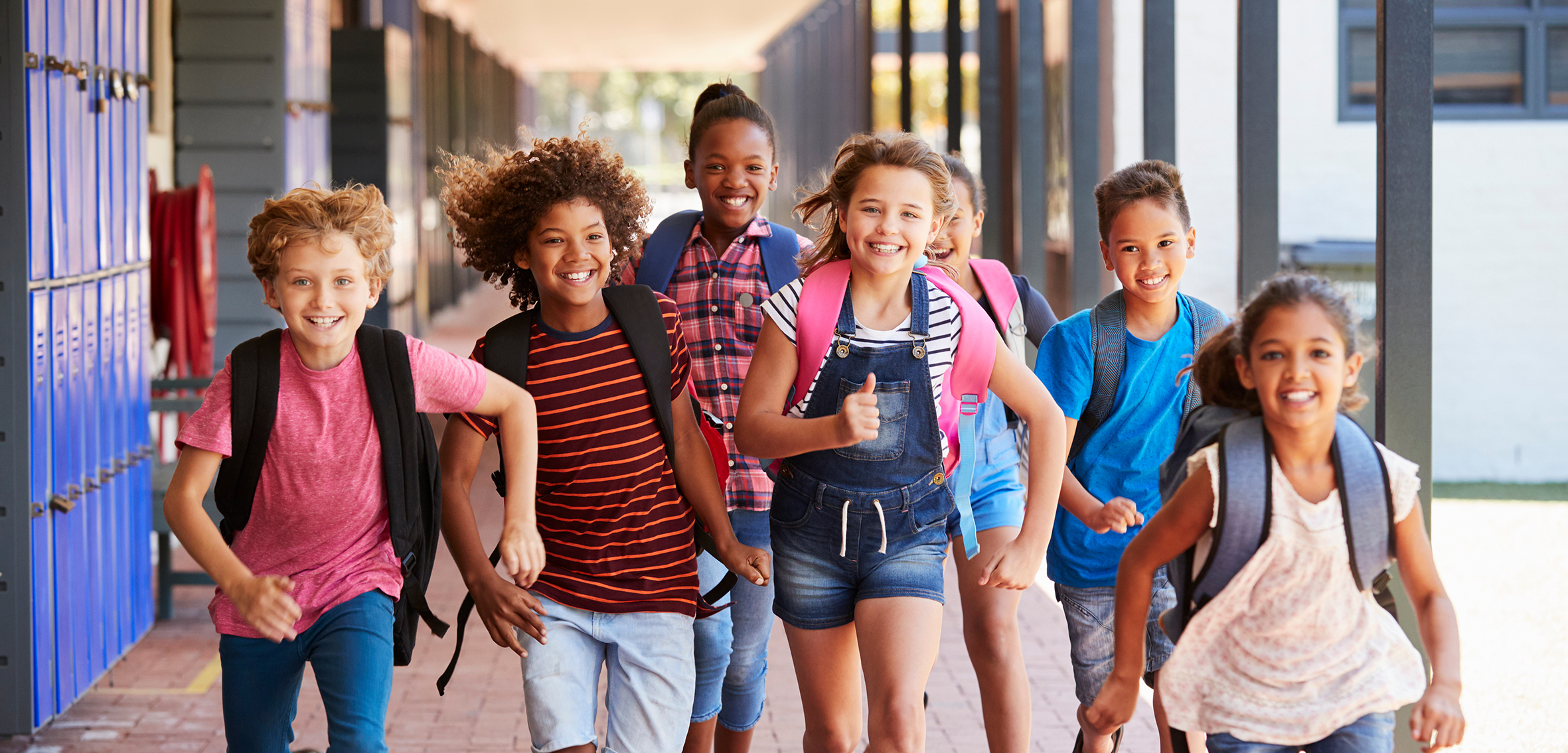 Gruppe von Grundschulkindern mit Schulranzen läuft lachend über den Schulhof vor einem Schulgebäude.