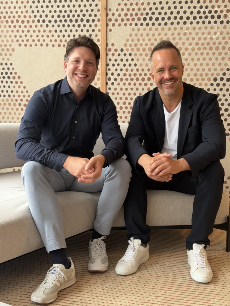Christian Nowak und Daniel Denker sitzen lächelnd auf einer Sofabank in einem modern gestalteten Innenraum und blicken in die Kamera.